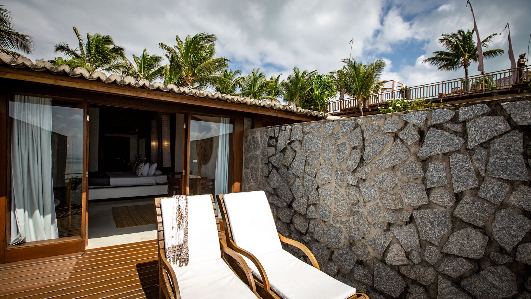 A stone-walled room with a bed, a wooden deck, and a view of palm trees.