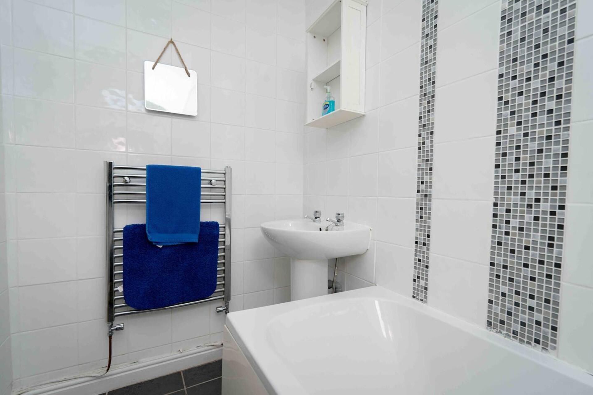 Un baño con un lavabo blanco, una toalla azul en un perchero y un mueble blanco sobre el lavabo, además de una zona de ducha con pared de azulejos.