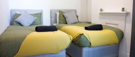 A room with two beds, each with a grey headboard and yellow bedding. There is a white fireplace with a small plant on the mantel.