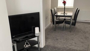 A living room with a black TV stand, a dining table with chairs, and a vase with flowers on the windowsill.