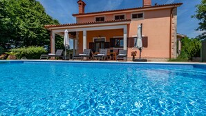 Ein Swimmingpool mit Liegestühlen und Sonnenschirmen, eine überdachte Terrasse und ein pfirsichfarbenes Haus mit Fensterläden.