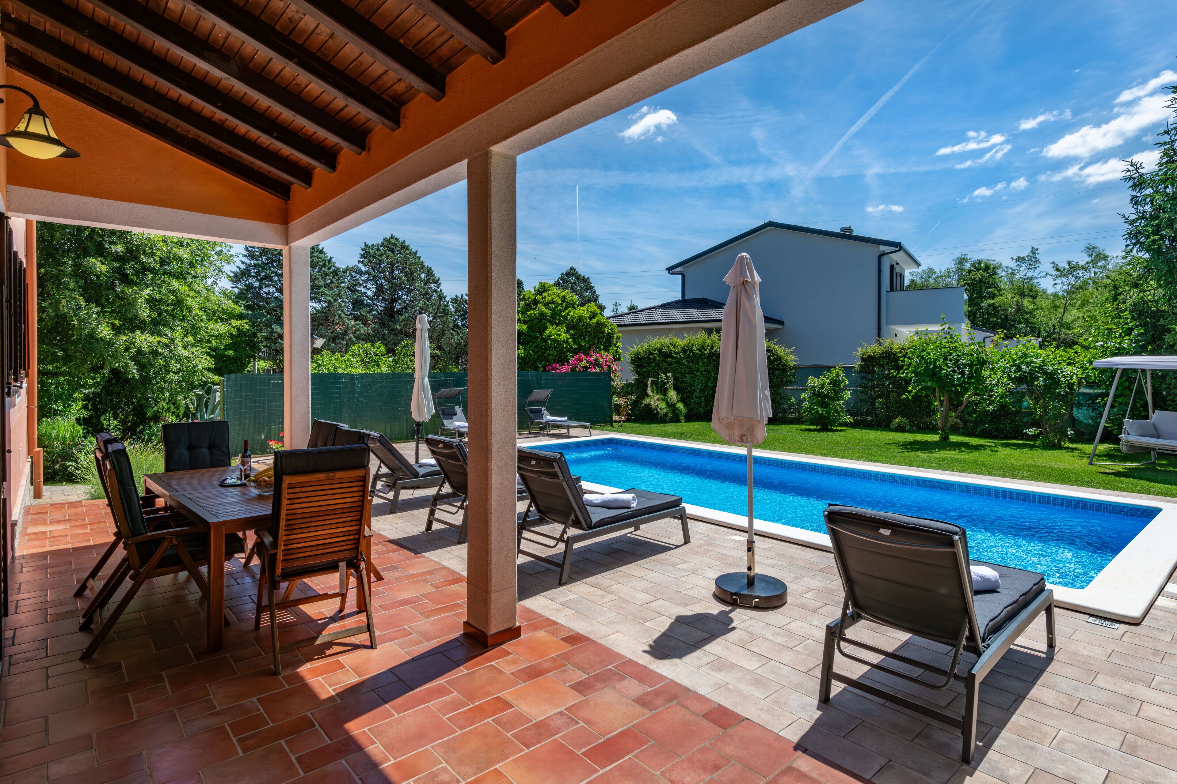 Un patio couvert avec une piscine, du mobilier d’extérieur et une vue sur une maison et la verdure.