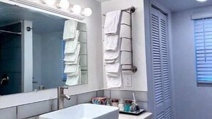 A bathroom with a white sink, vanity, and a mirror with lights. There are towels hanging on a rack and a window with blinds.