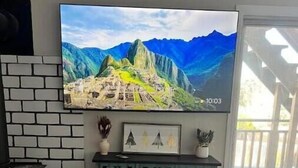 A living room with a large flat-screen TV mounted on the wall displaying a mountain landscape. There is a decorative cabinet with intricate carvings below the TV.