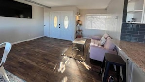 A spacious living area with a dark wooden floor, a white sofa, and a small table. There is a set of double doors with glass panels and a window with blinds.