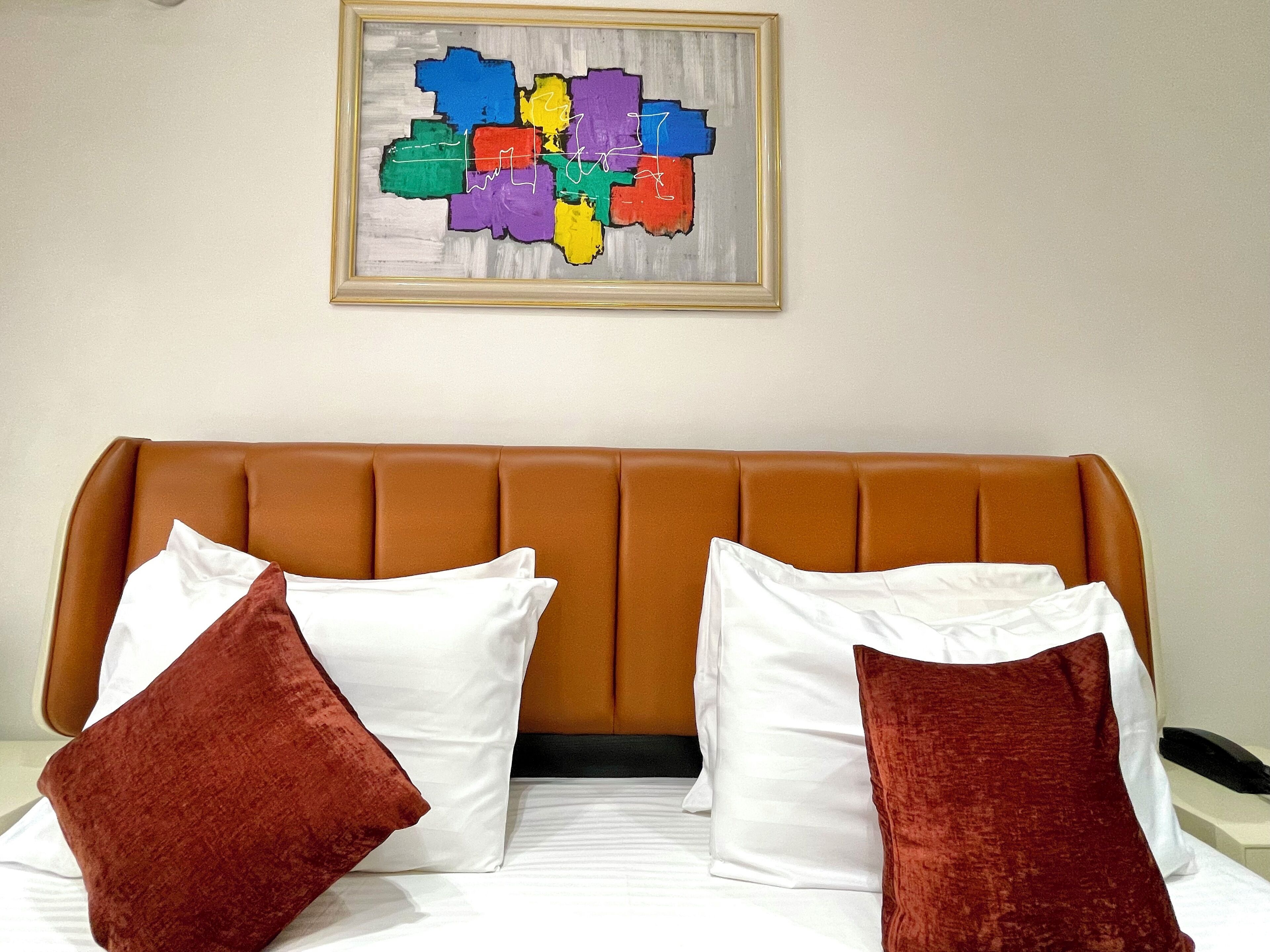 Een hotelkamer met een bruin gestoffeerd hoofdeinde, witte beddengoed en twee rode kussens.