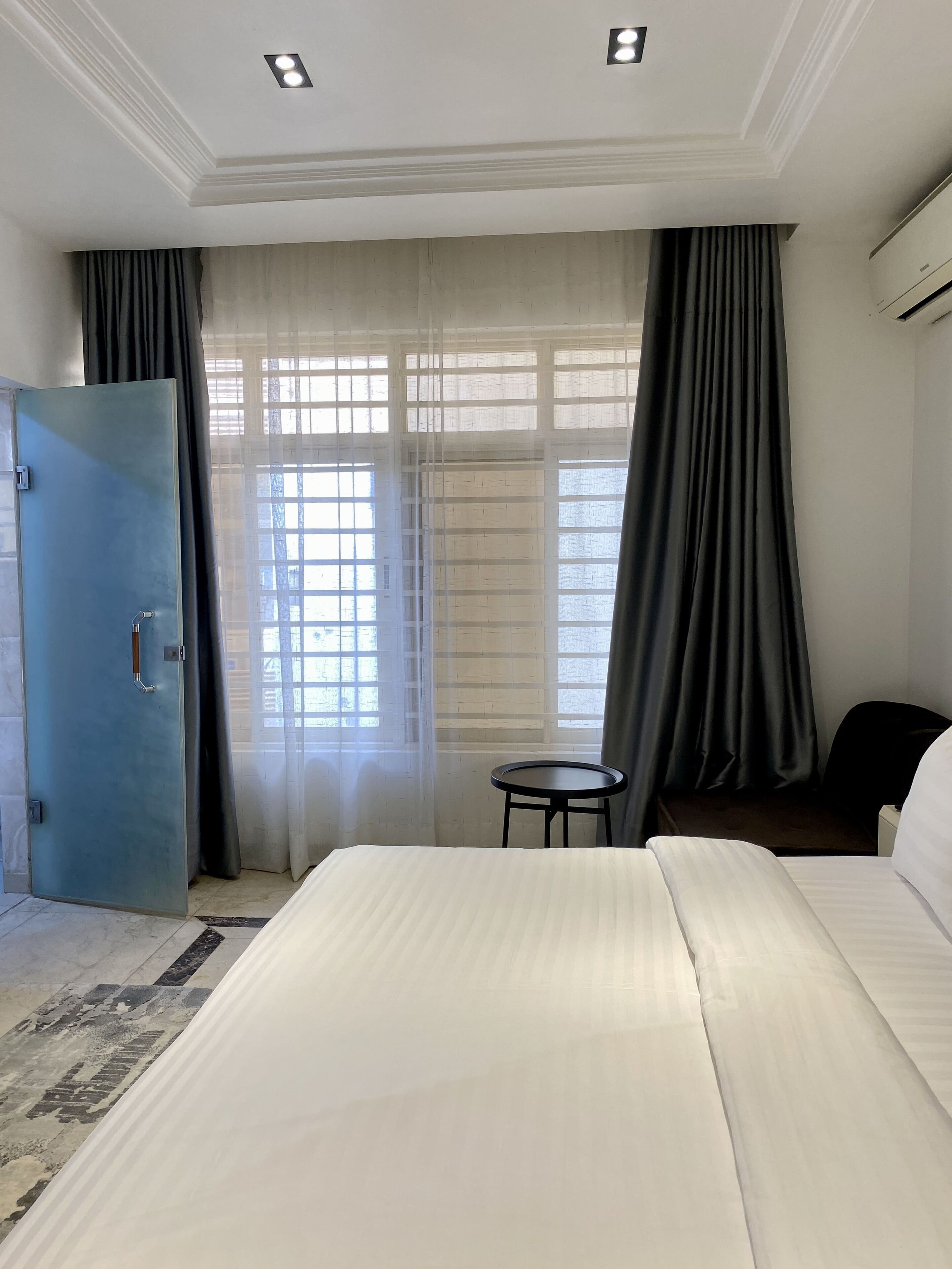 Quarto de hotel com cama, mesa redonda pequena, cadeira, guarda-roupa e janela com cortinas.