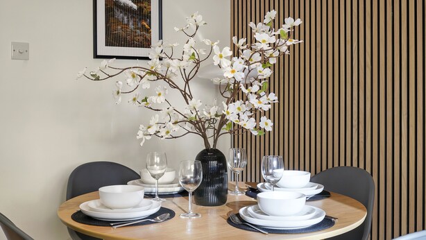 Ein Esstisch ist mit weißen Tellern, Schalen und Weingläsern gedeckt, dazu steht eine Vase mit weißen Blumen.