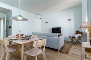 A modern living room with a grey sofa, wooden dining table, and chairs. A flat-screen TV is mounted on the wall, and there is a lamp on a side table. - Modern & Cozy Faro Mercado Apt 21 by Lisbeyond (Faro)
