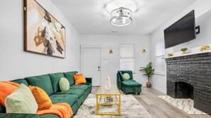 Un salon avec un canapé vert, un petit fauteuil vert, une table basse en marbre, une cheminée noire et un téléviseur fixé au mur.