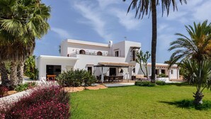 A modern two-story building with a white facade, a covered patio area, and a well-maintained lawn with tropical plants. - CAN COSMI (Santa Eulalia del Río)