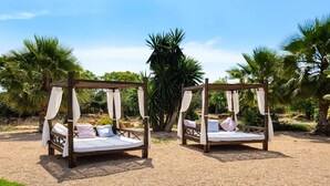 Two outdoor wooden beds with white bedding and curtains, set on a gravel path with palm trees in the background. - CAN COSMI (Santa Eulalia del Río)