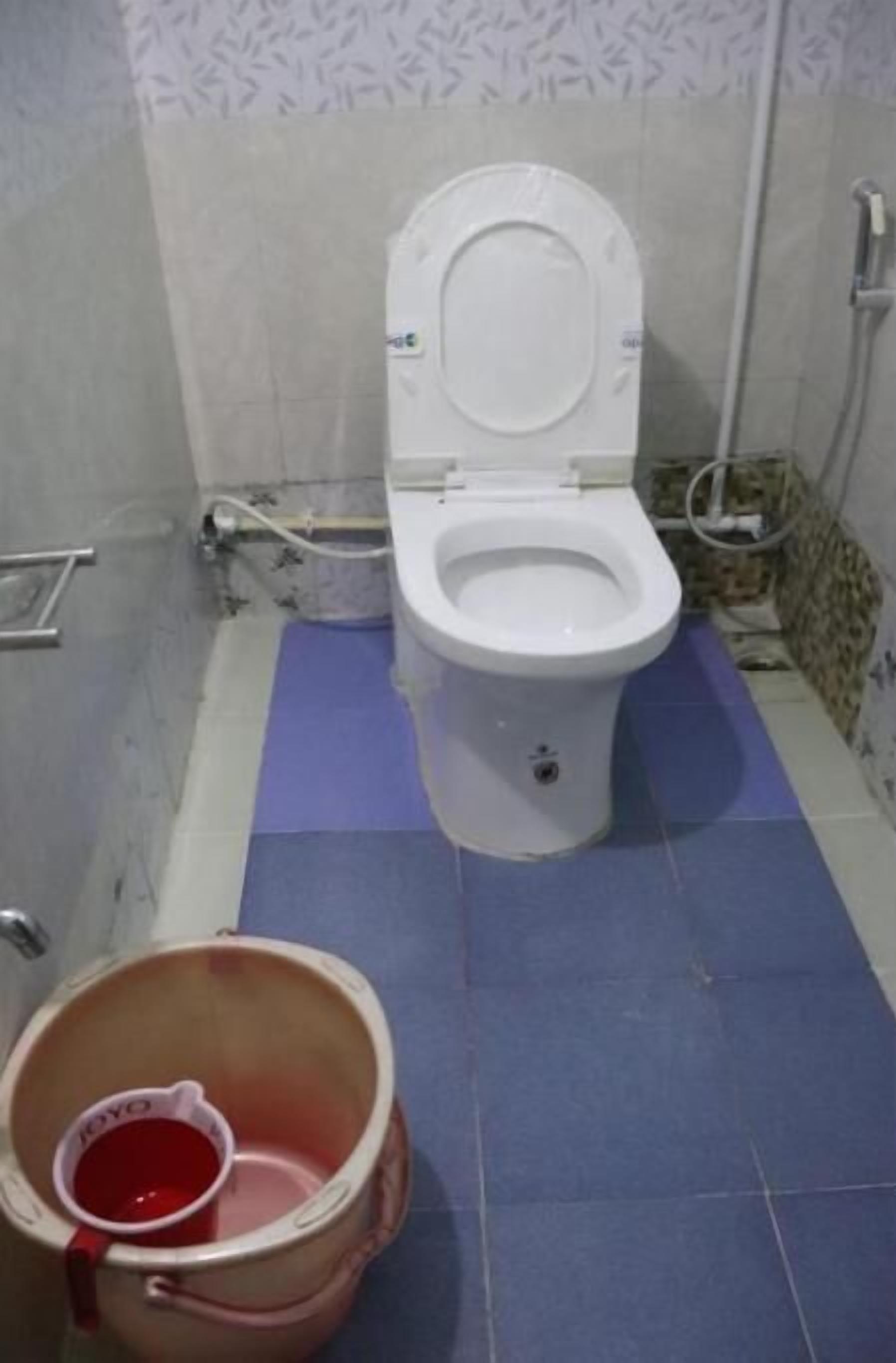 Une salle de bain avec des toilettes blanches, un tapis de sol bleu et un seau contenant un liquide rouge.