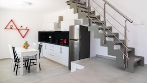 Una cocina moderna con refrigerador de acero inoxidable, armarios blancos y encimera negra. Hay una mesa de comedor con sillas y un adorno mural rojo de diseño geométrico. Una escalera original con barandilla de metal conduce a otro nivel.
