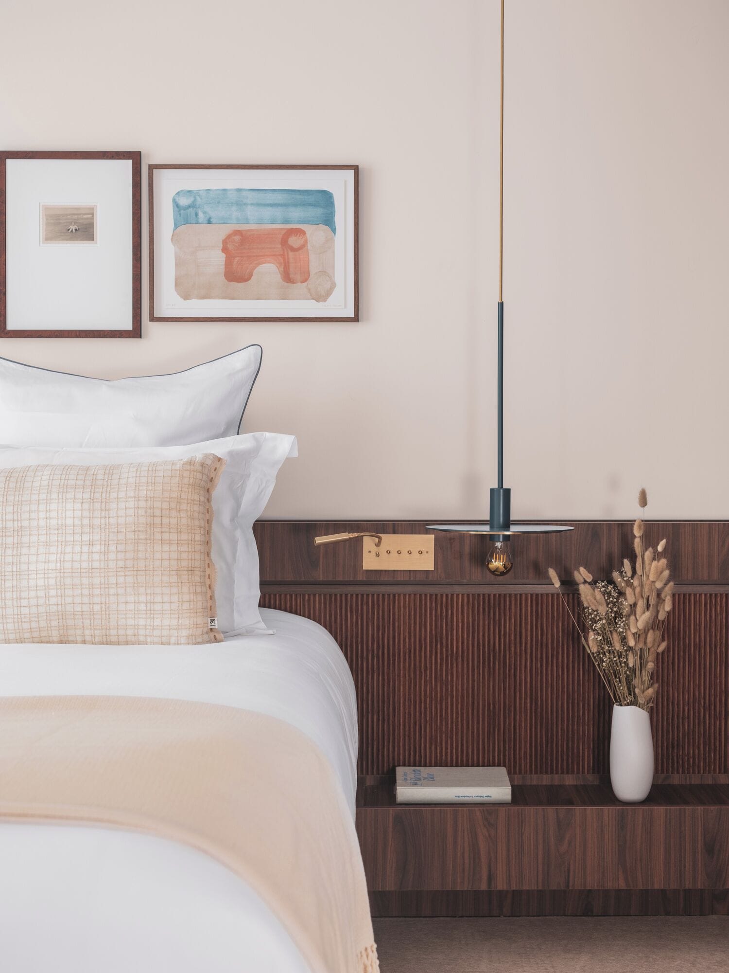 Un lit bien fait, avec une literie blanche et beige, une table de chevet avec une lampe, et deux œuvres d’art encadrées accrochées au mur.