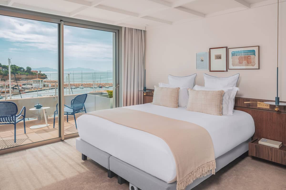 Habitación de hotel con una cama grande, un balcón con vistas al mar y una pequeña mesa con sillas.