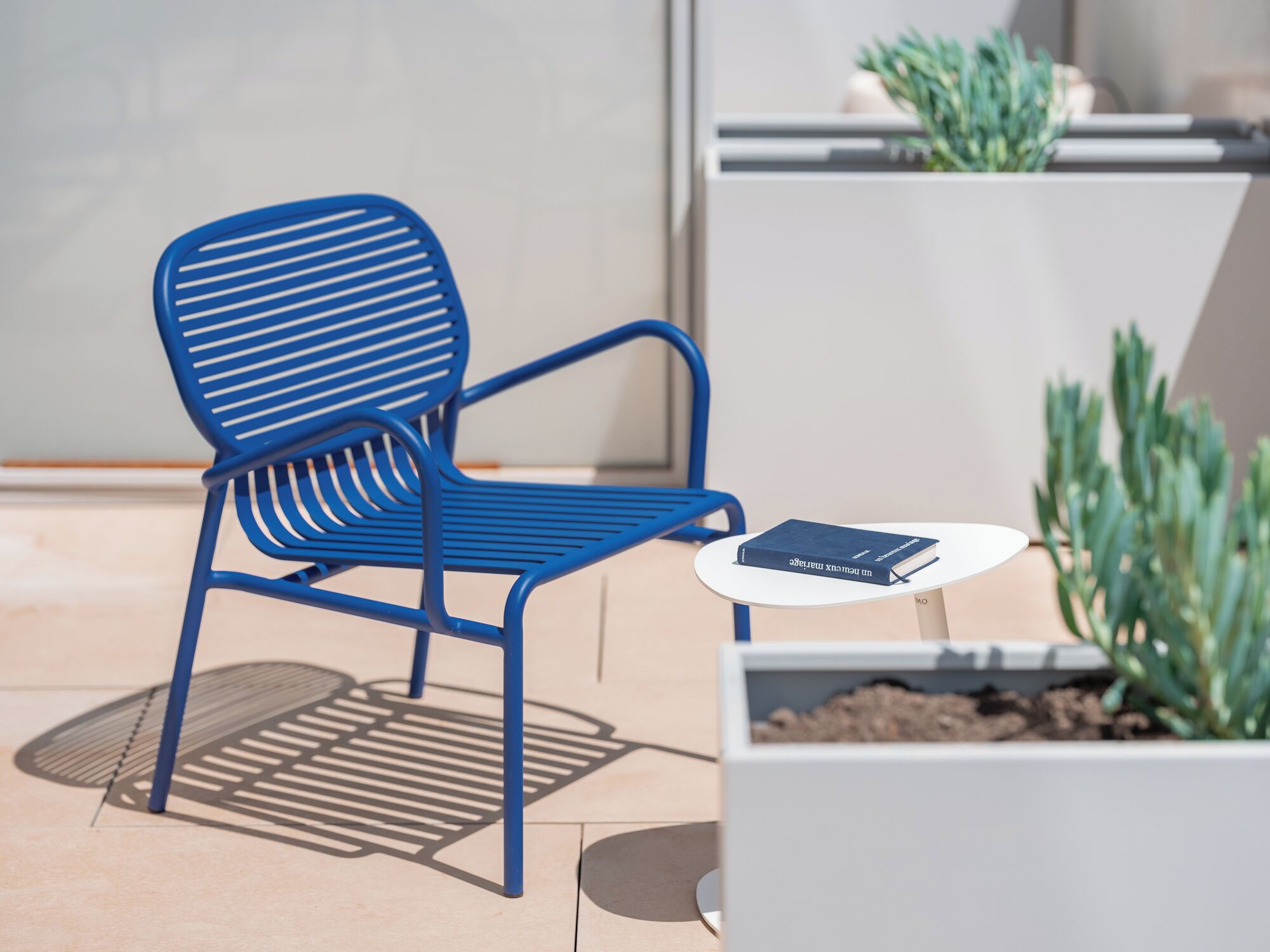 A blue metal chair with a slatted backrest and armrests, positioned next to a small white table with a book on it.