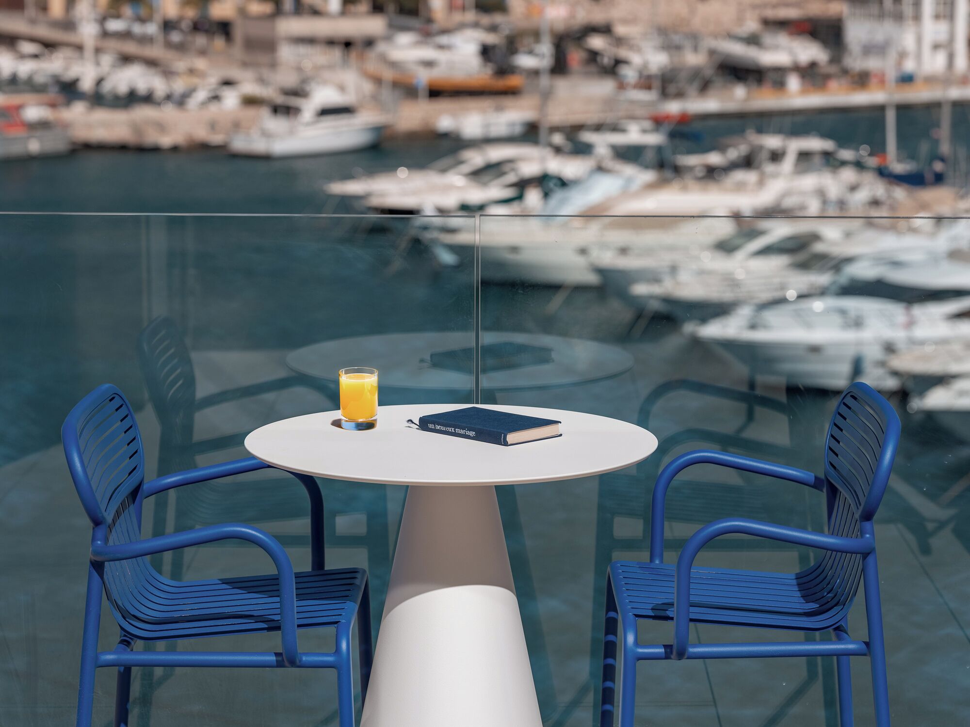 Et glasbord med to blå stole, et glas appelsinjuice og en bog med titlen "Um Beaux Mariage" på en balkon med udsigt over en havn med både.
