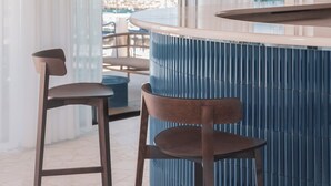 Zona de bar moderna amb un taulell corbat, dos tamborets de fusta i un gran finestral amb vista a una platja.