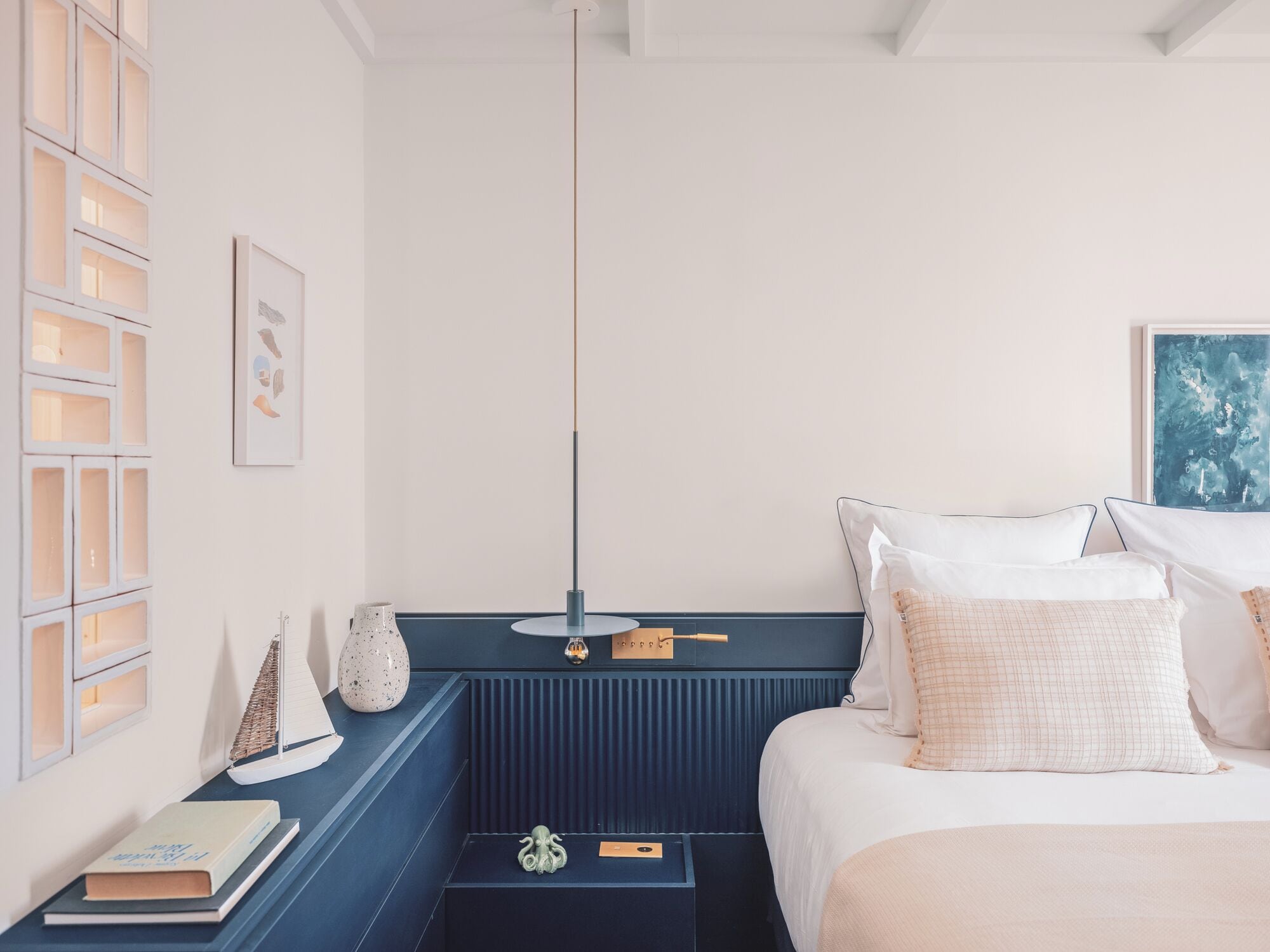 A bedroom with a bed, bedside tables, a hanging light, a wall-mounted shelf, and a framed picture.