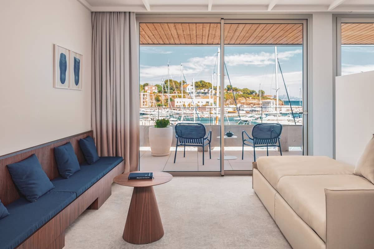 Una sala de estar moderna con un sofá, una mesa de centro y dos sillones, con vistas a un puerto deportivo con barcos y edificios.