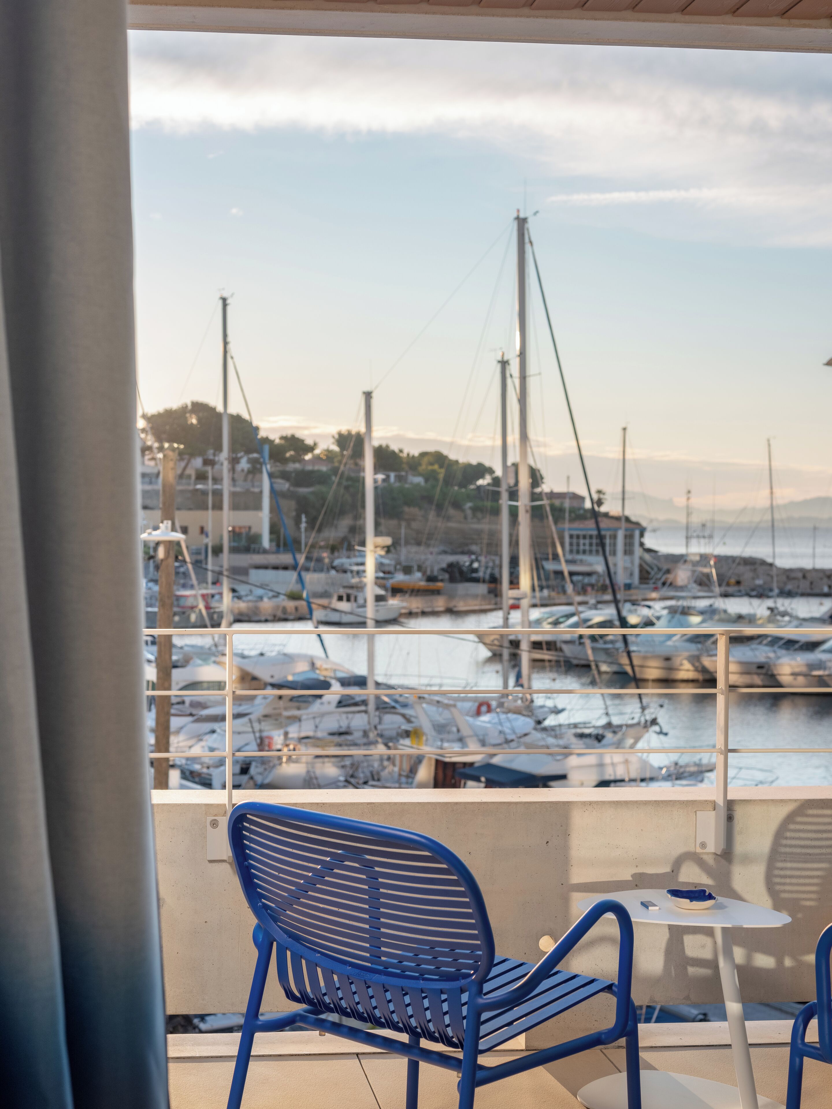 A view from a balcony with a blue chair and a small table, overlooking a marina with boats and a distant shoreline.