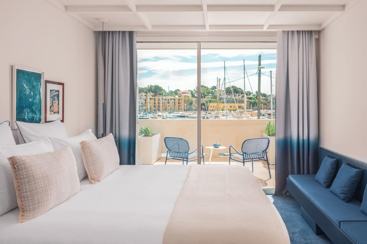 Habitación de hotel con una cama grande, un sofá, un balcón con sillas y vistas a barcos y edificios.