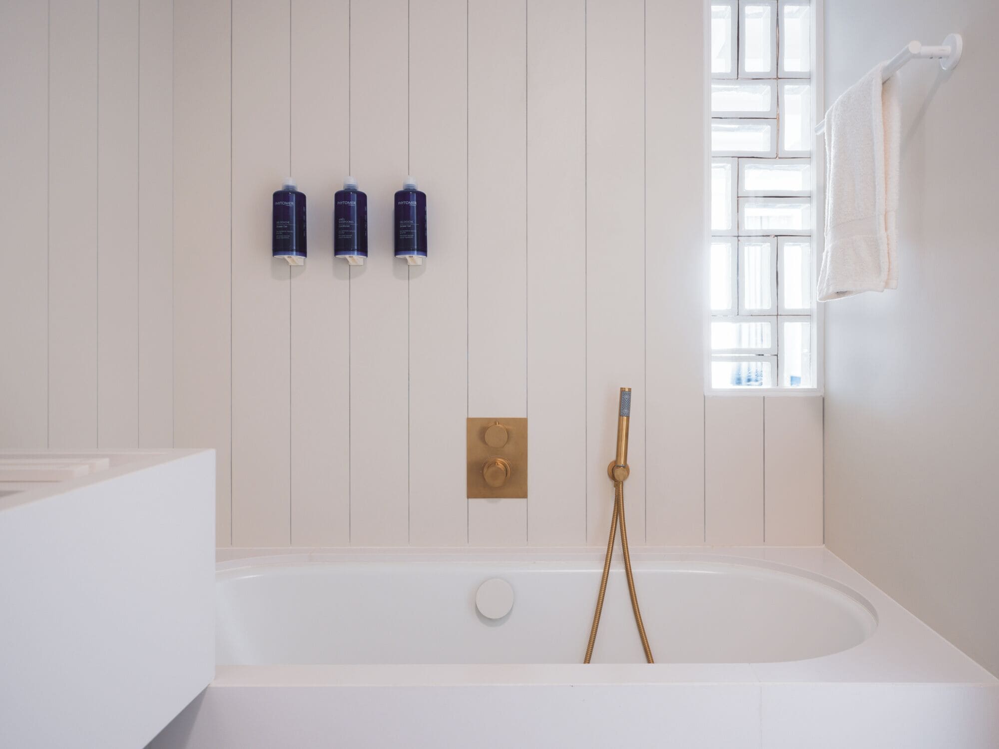 A modern bathroom with a white bathtub, a gold showerhead, and three bottles of shampoo on the wall.