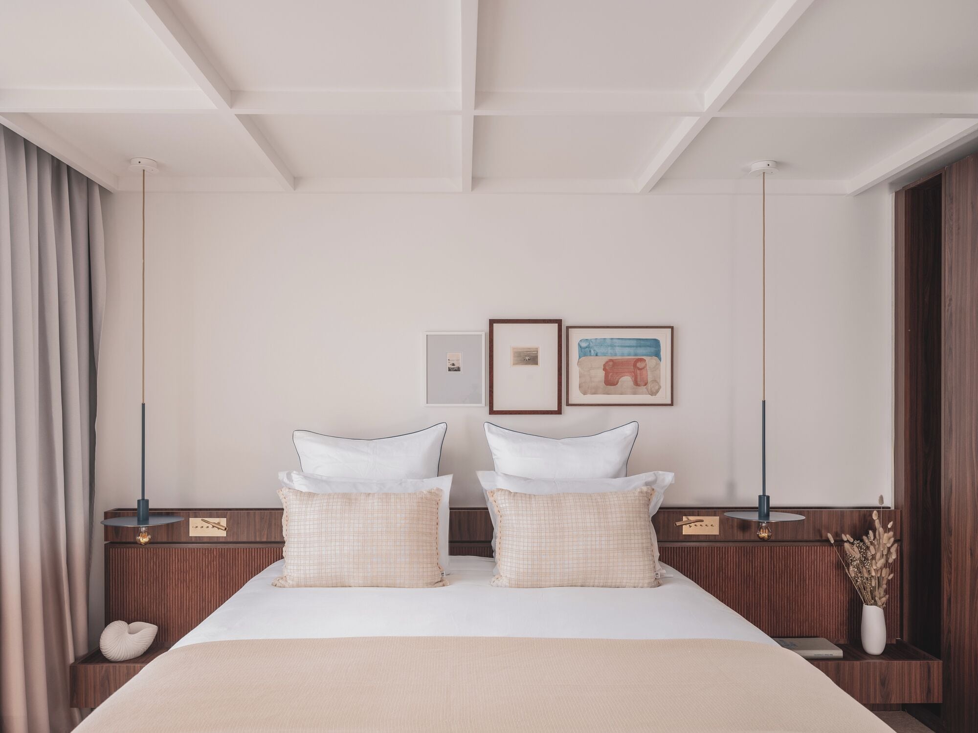 Une chambre d’hôtel moderne avec un grand lit, deux tables de chevet, une lampe fixée au mur et un tableau encadré.