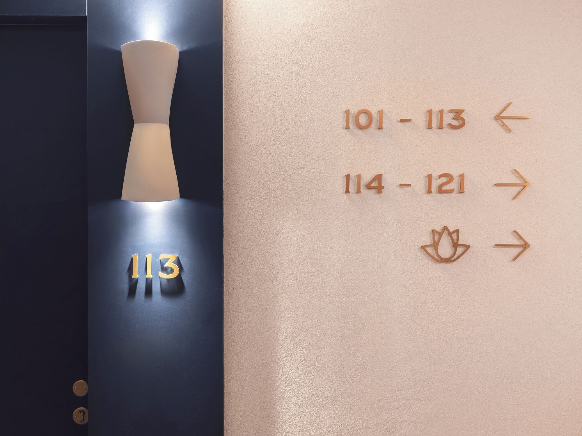 Une chambre d’hôtel avec une porte bleu foncé portant le numéro 113. Des panneaux indicateurs sont fixés au mur, indiquant les numéros des chambres et les directions.