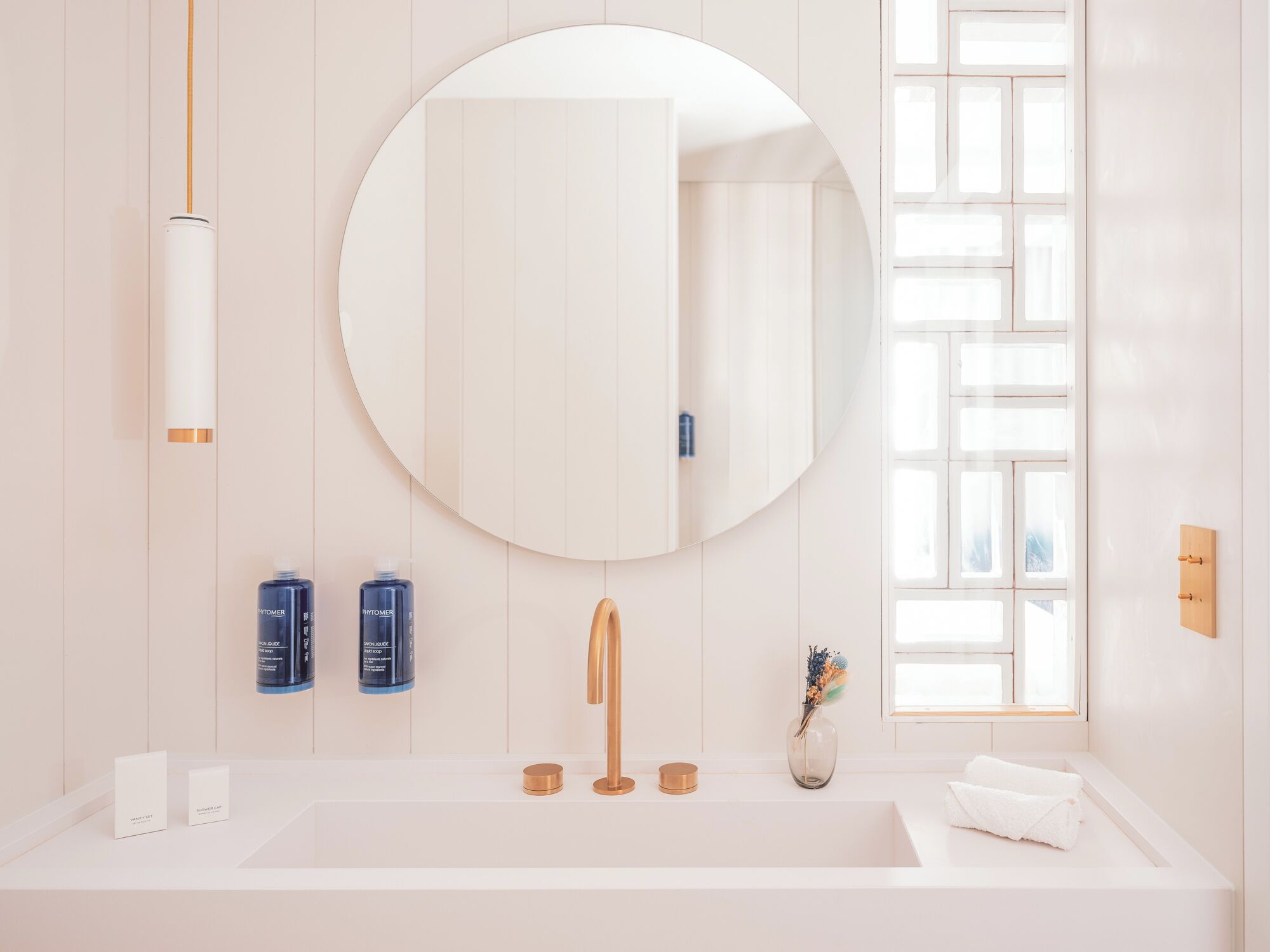 A modern bathroom with a large round mirror, a white sink, and two blue bottles on the counter.