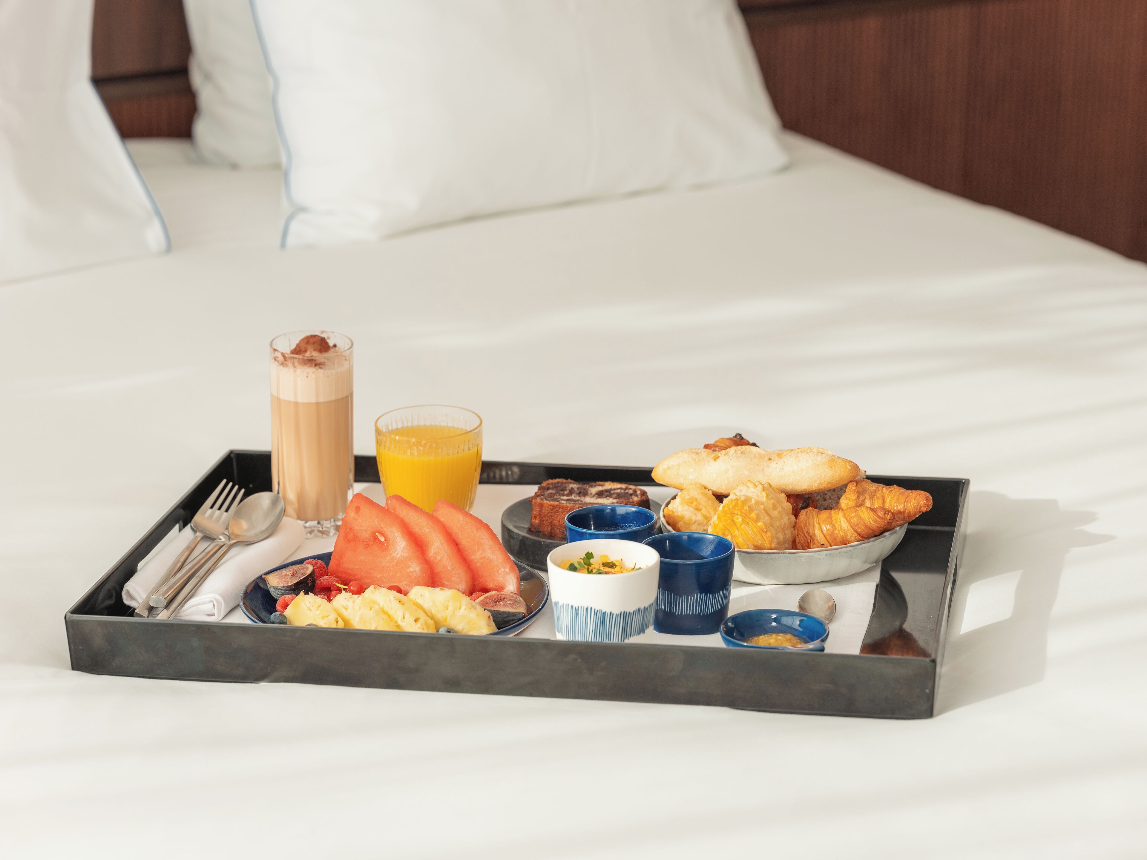 Bandeja de desayuno con croissants, fruta y bebidas sobre la cama.