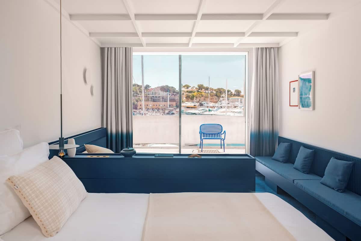 Una habitación de hotel moderna con una cama grande, un sofá azul, una silla azul y vistas a un puerto deportivo.