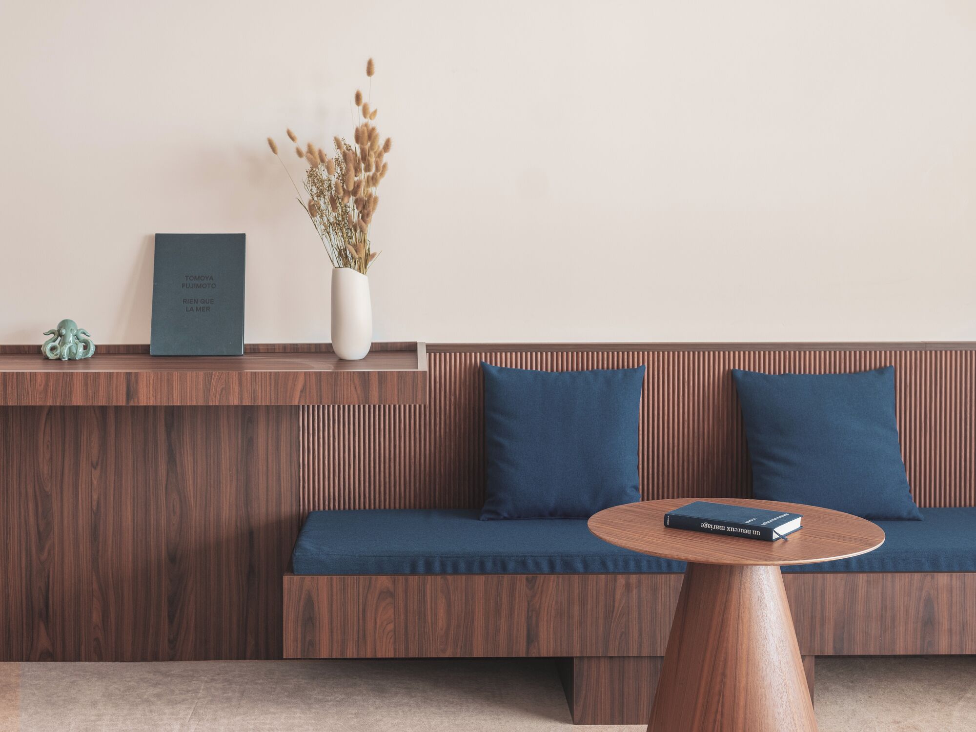 Um interior moderno com uma parede de painel de madeira, um sofá azul com almofadas, uma mesa de centro redonda de madeira, um vaso branco com plantas secas e um pequeno objeto decorativo na estante.