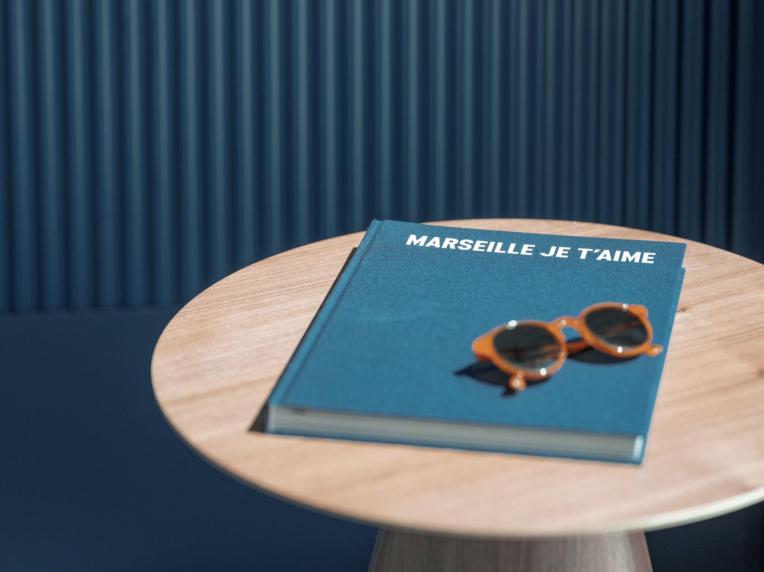 Ein Buch mit dem Titel „MARSEILLE JE T’AIME“ auf einem runden Holztisch, mit einer Sonnenbrille darauf.