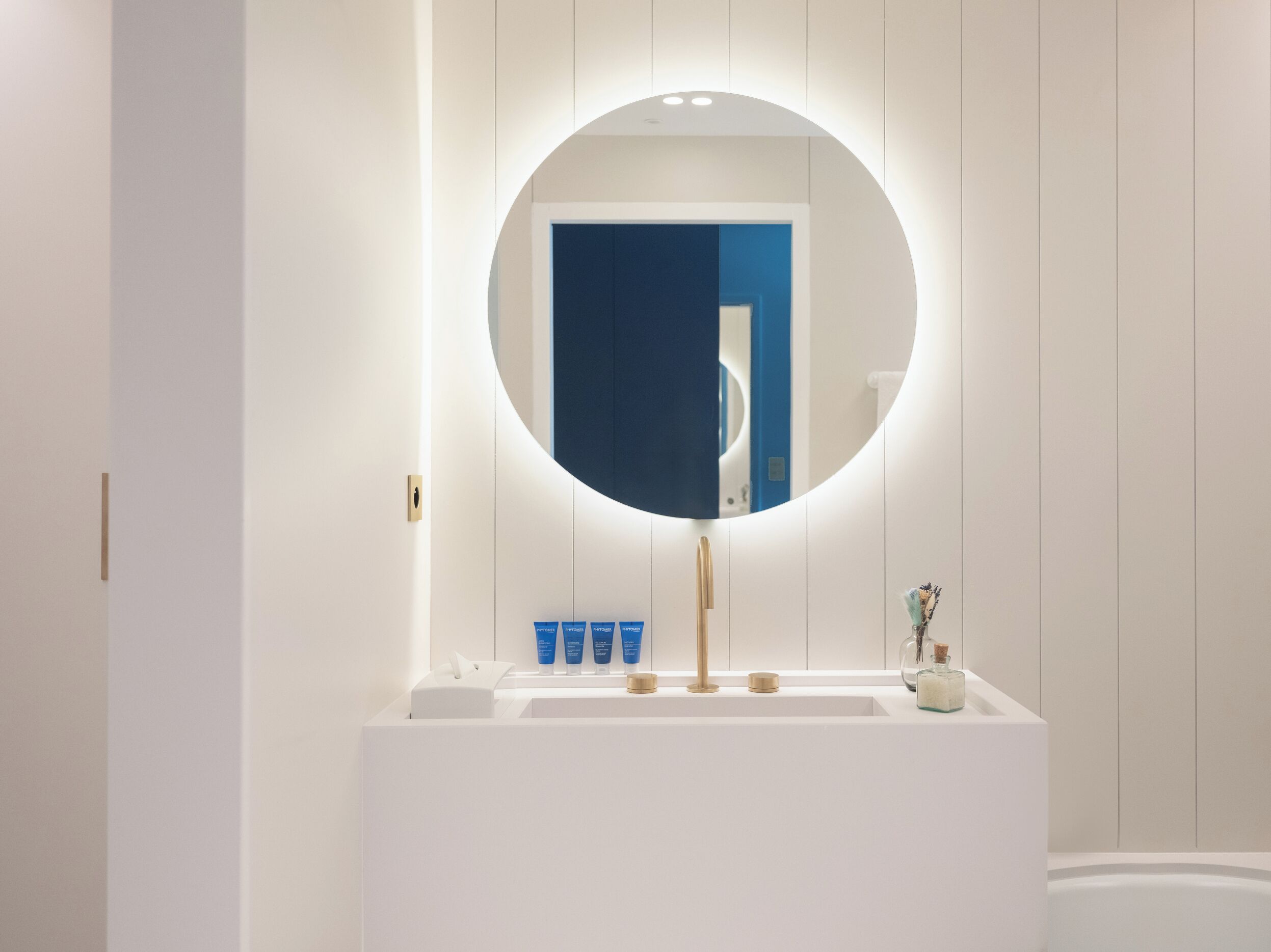 Ein modernes Badezimmer mit einem weißen Waschtisch, einem runden Spiegel mit Beleuchtung und einer blauen Tür im Hintergrund.