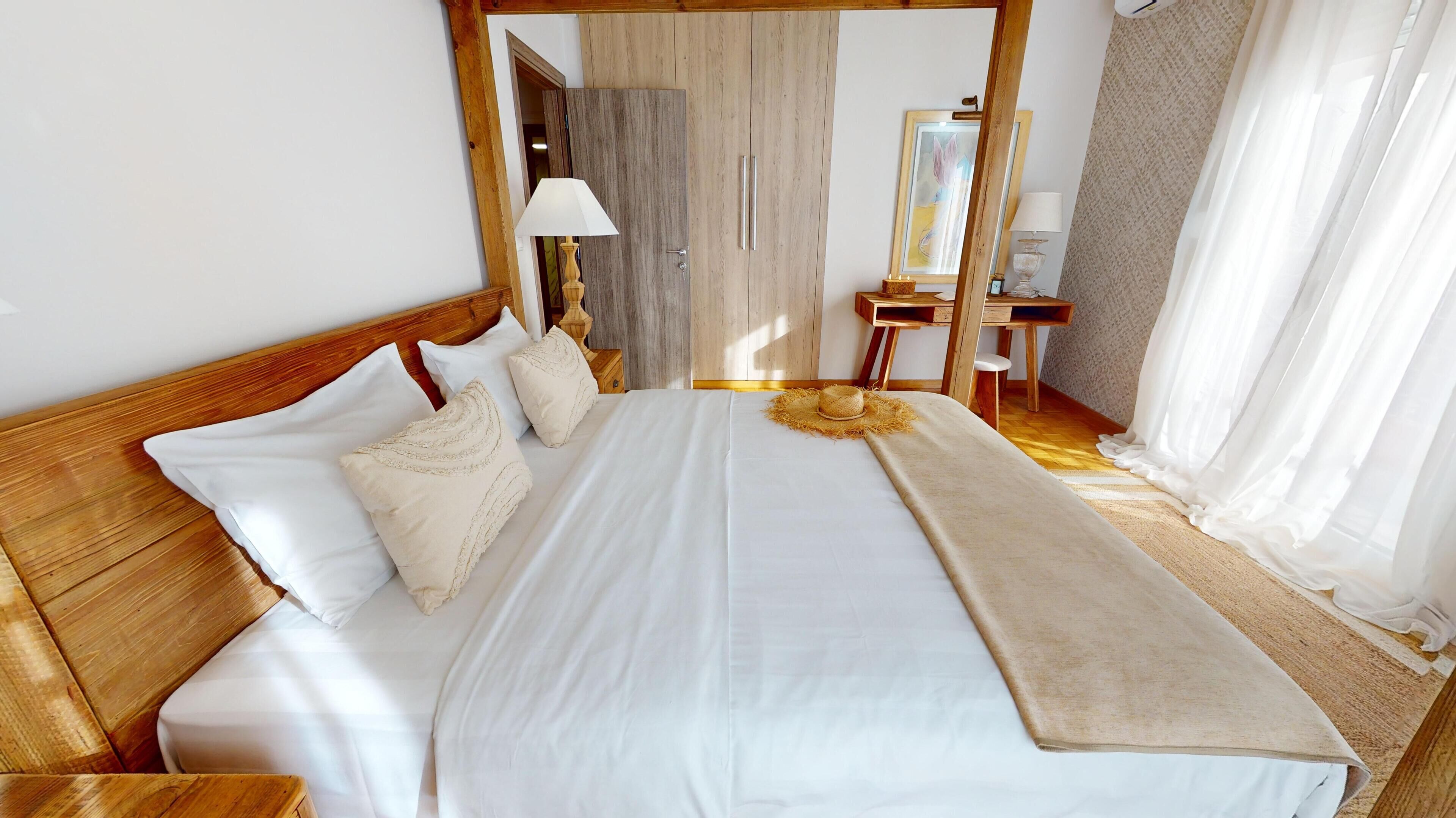 A neatly made bed with white linens and a beige throw blanket, a wooden headboard, and a wooden nightstand with a lamp.
