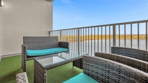 En balkon med fletmøbler, et glasbord og udsigt over et vandareal og et fjerntliggende landskab.