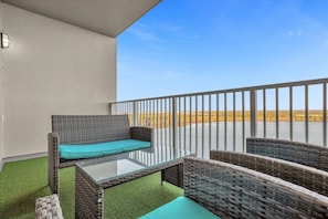 A balcony with wicker furniture, a glass table, and a view of a water body and distant landscape.