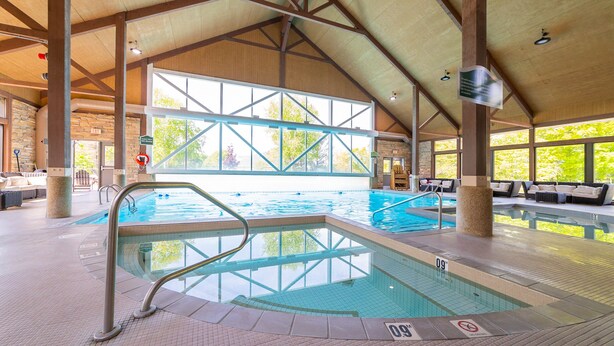 An indoor swimming pool with clear glass walls, a tiled floor, and a high ceiling with wooden beams. - Horseshoe Valley Suites - The Glade (Oro-Medonte)