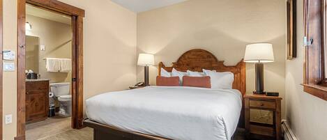 Habitación de hotel con cama, dos mesitas de noche con lámparas, ventilador de techo, cabecero de madera, armario de madera, baño con inodoro y espejo.