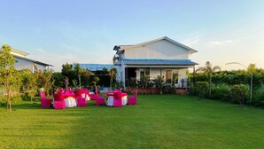 Un espace extérieur verdoyant, avec des chaises roses disposées en rangées, un bâtiment blanc surmonté d’un toit bleu et un ciel dégagé.