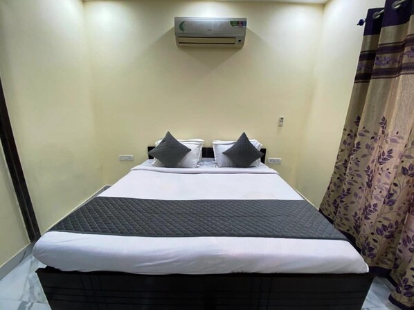 A single bed with a black quilted headboard, white bedding, and two dark pillows. An air conditioning unit is mounted on the wall above the bed.
