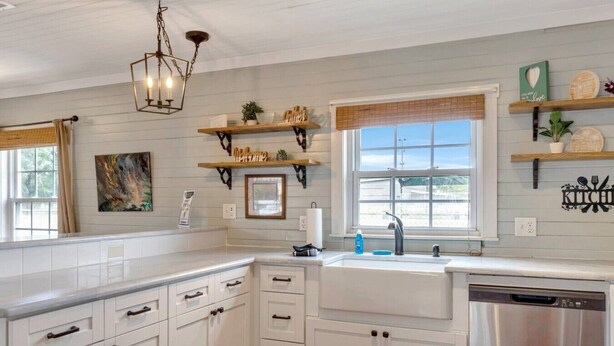Cocina con alacenas blancas, lavavajillas, ventana con persianas y objetos decorativos en estantes.