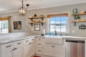 Cocina con alacenas blancas, lavavajillas, ventana con persianas y objetos decorativos en estantes.