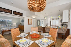 A dining area with a round table set for four, surrounded by patterned chairs, a kitchen in the background, and a bedroom with a bed and window blinds.