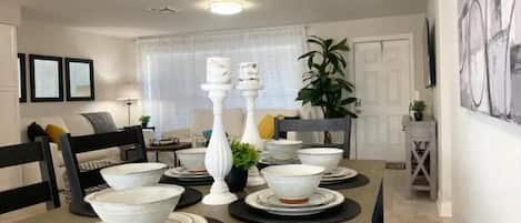A dining area with a wooden table set with white bowls and plates, surrounded by black placemats. There are chairs around the table, and a plant is visible in the background.