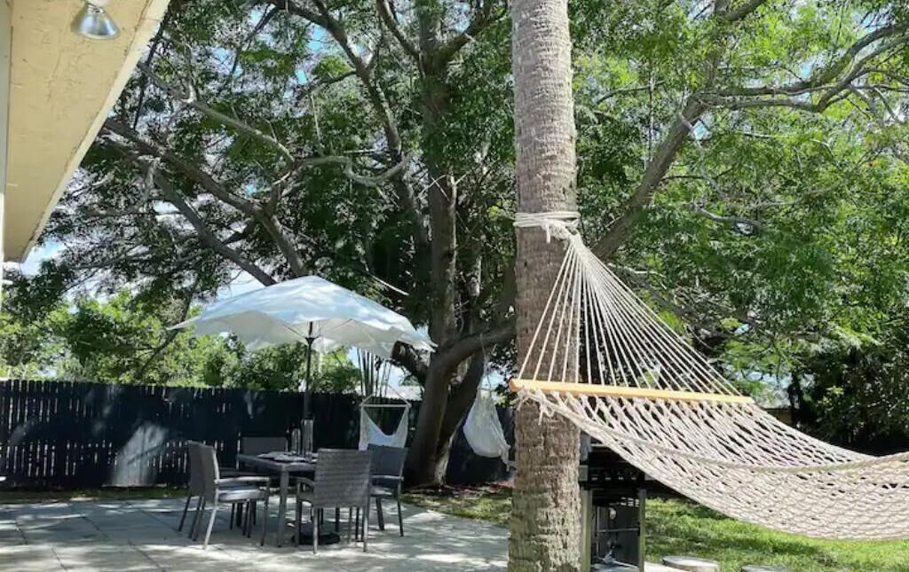 A hammock tied to a tree in a backyard with outdoor furniture and umbrellas.