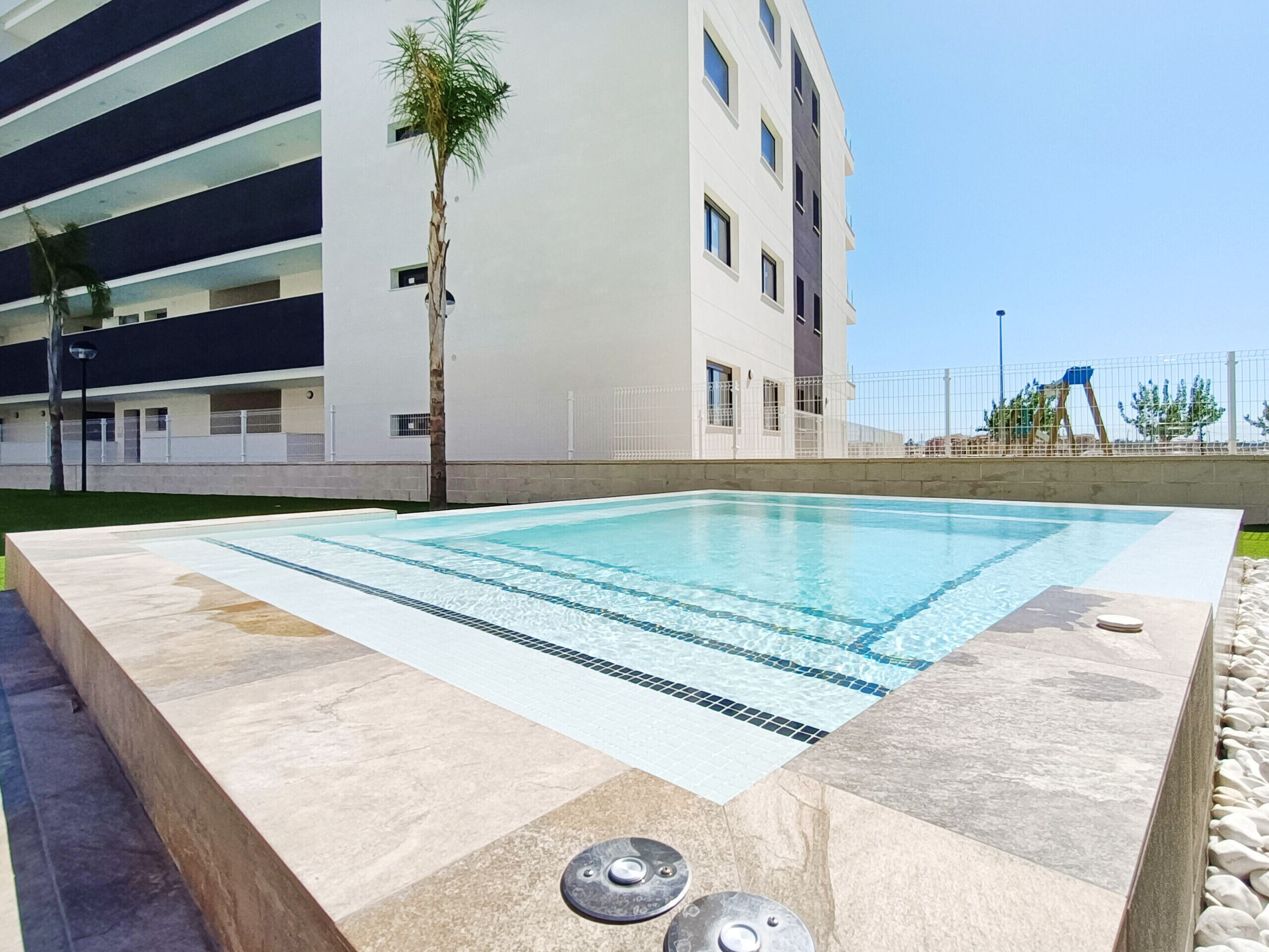 Una piscina rectangular con borde de azulejos, rodeada de baldosas de piedra, frente a un edificio moderno con varios balcones y una palmera.