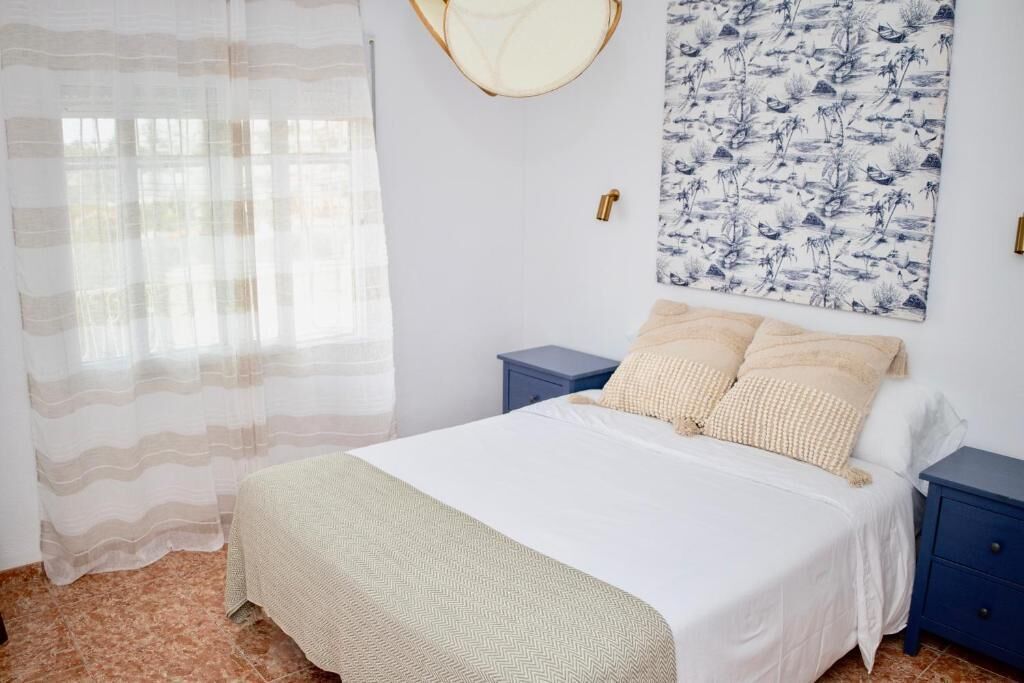 A bedroom with a bed, a nightstand, a wall-mounted lamp, a window with sheer curtains, and a floral-patterned wall hanging.